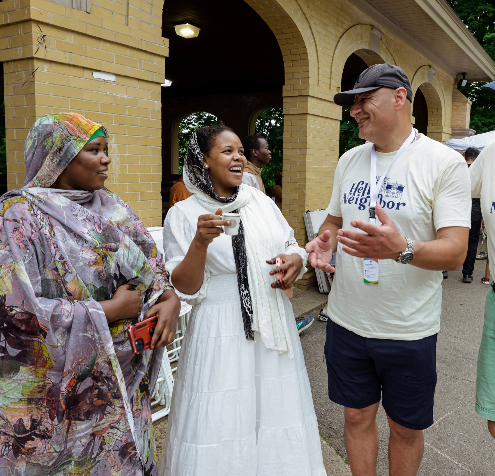 Two Hello Neighbor volunteers in branded T-shirts laugh mid-conversation with two women holding coffee at an outdoor summer gathering.
